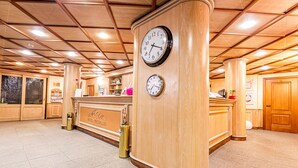 A reception area with wooden paneling, two wall clocks, a reception desk, and a tiled floor. - Hotel Cristallo (Sestriere)