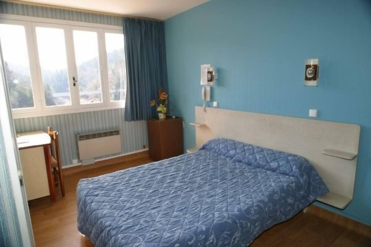 A bedroom with a bed, a desk, a chair, a dresser, a window with curtains, and a wall-mounted telephone.