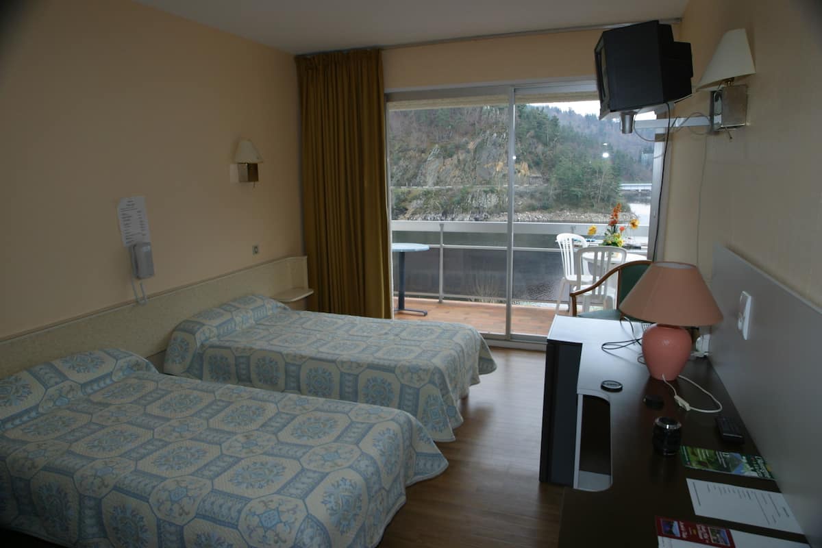 A hotel room with two beds, a desk with a lamp, a TV, and a view of a balcony with a chair and flowers.