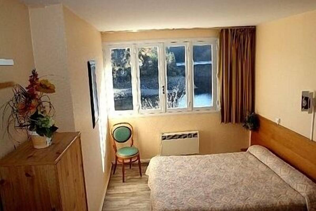 A small bedroom with a bed, a chair, a radiator, and a window with curtains.