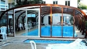 A covered outdoor swimming pool with clear glass walls, surrounded by white plastic chairs and tables. - Garabit Hôtel (Anglards-de-Saint-Flour)