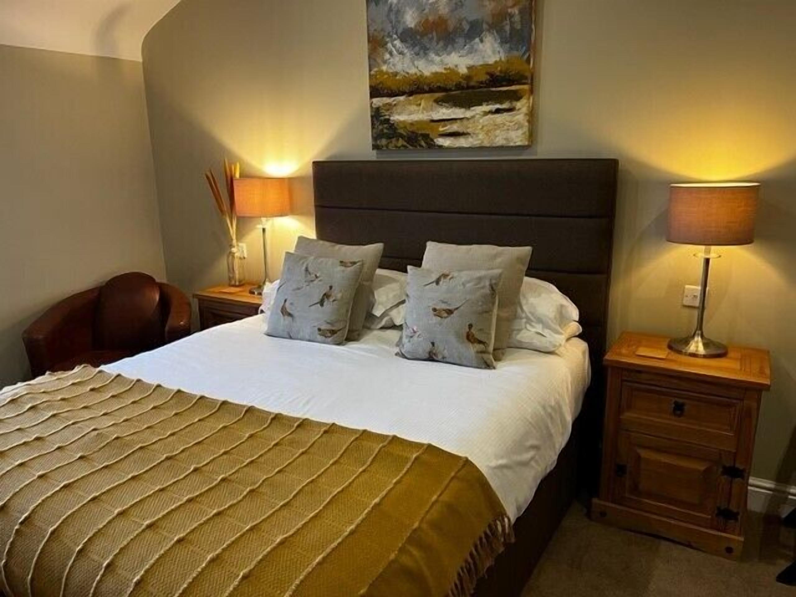 A neatly made bed with a patterned throw, two bedside lamps, a painting above the bed, and a wooden nightstand with drawers.
