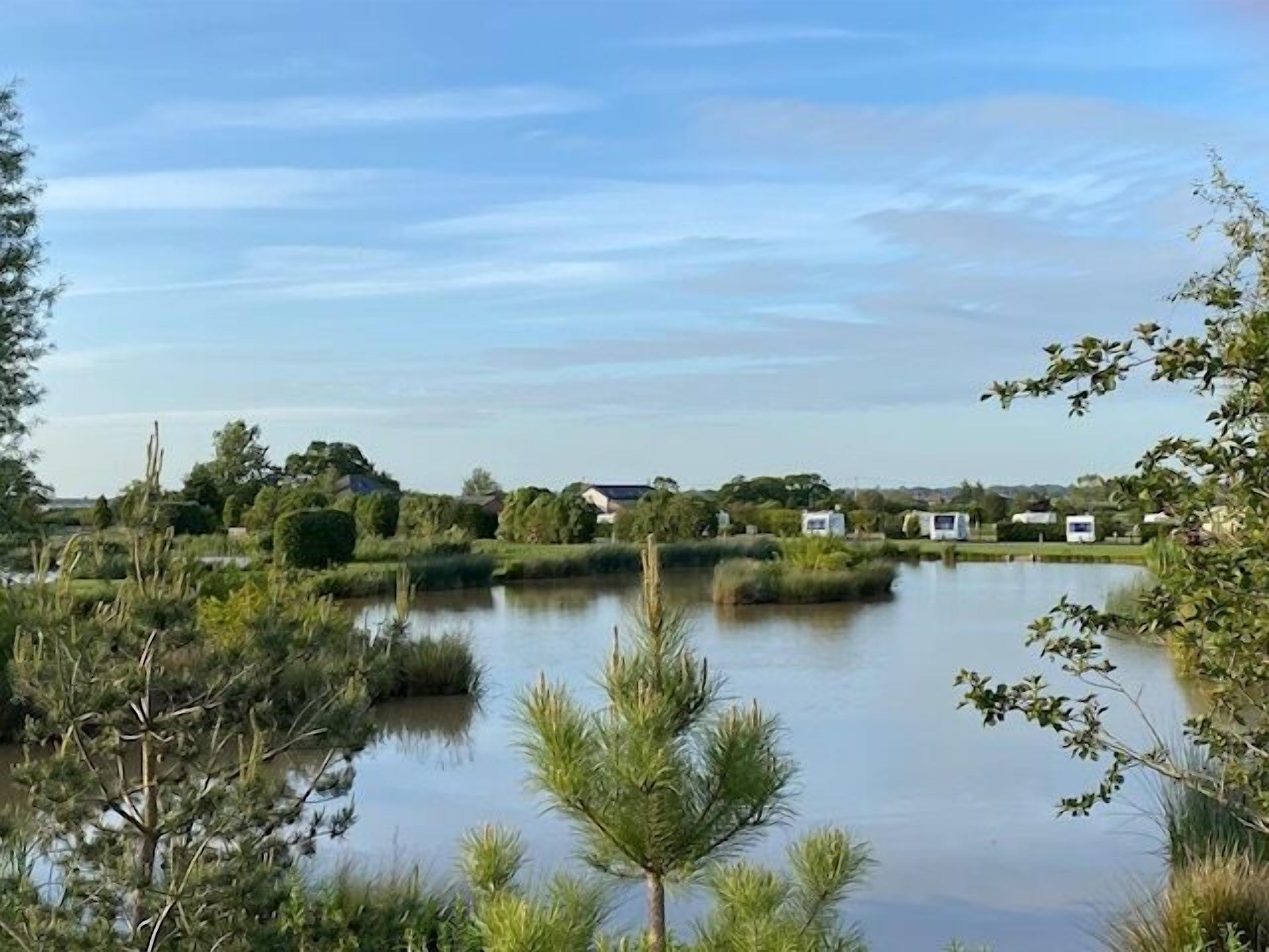 A serene lakeside view with a calm water body, surrounded by lush greenery and a few buildings in the background.
