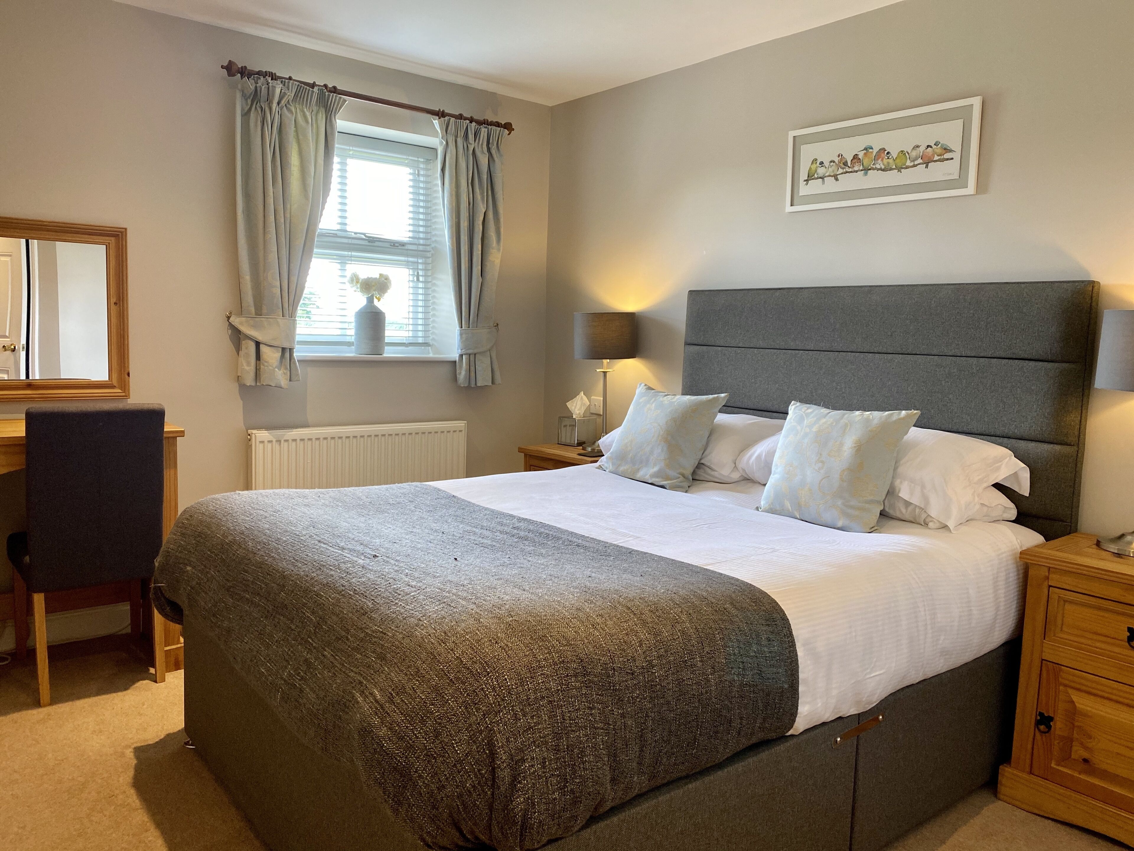A neatly arranged bedroom with a large bed, a bedside table with a lamp, a chair, a window with curtains, and a wall-mounted picture.