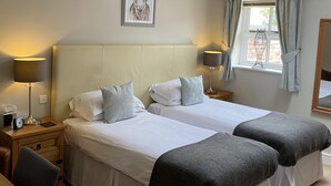 A hotel room with two beds, a desk, a chair, a bedside table with a lamp, a framed picture on the wall, and a window with curtains. - New Farm Bed & Breakfast (Winsford)