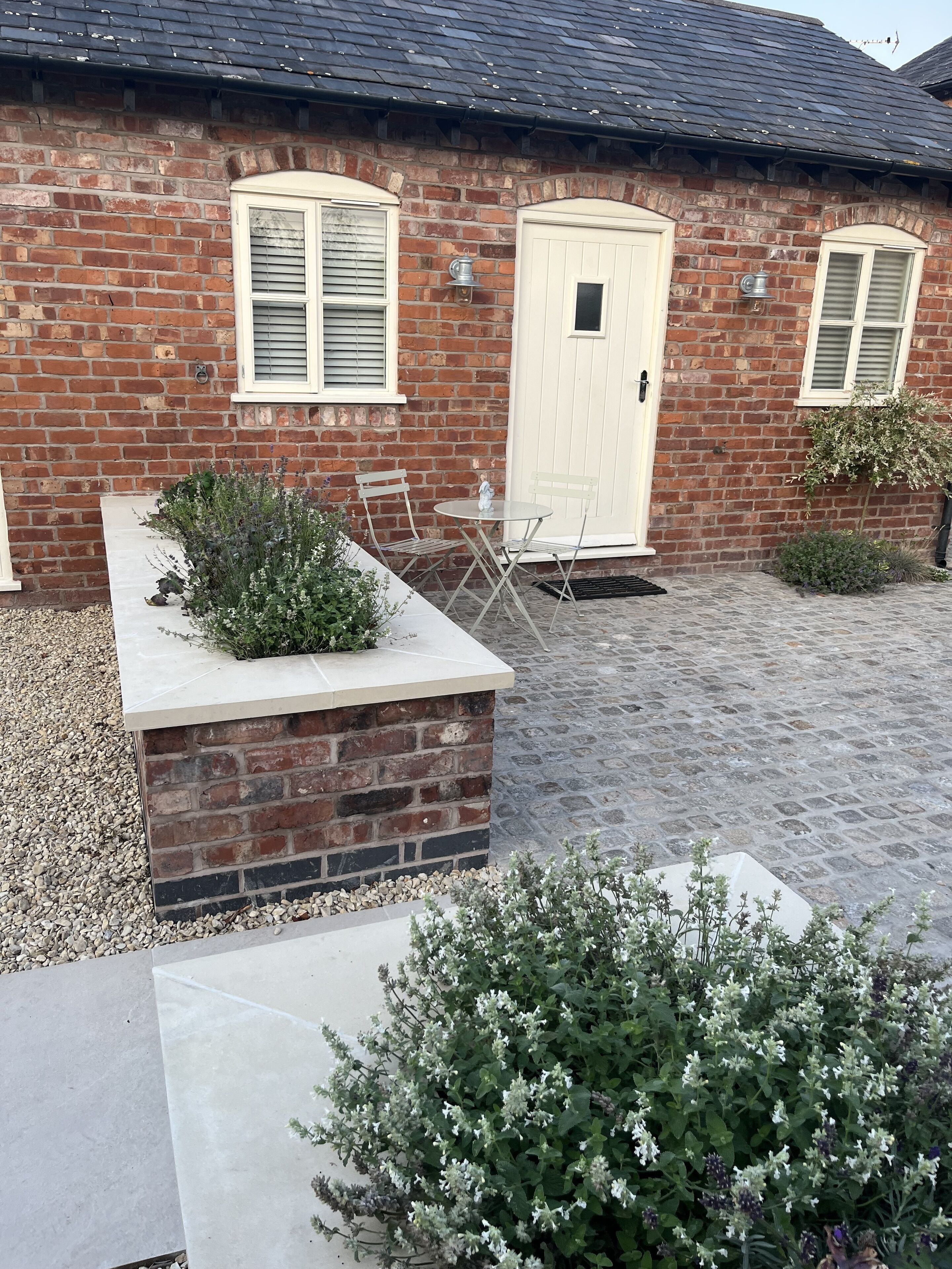 Une maison en briques avec une porte blanche, un petit patio avec une table et des chaises, et un jardin agrémenté de différentes plantes.