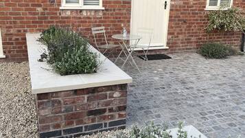 Une maison en briques avec une porte blanche, un petit patio avec une table et des chaises, et un jardin agrémenté de différentes plantes.