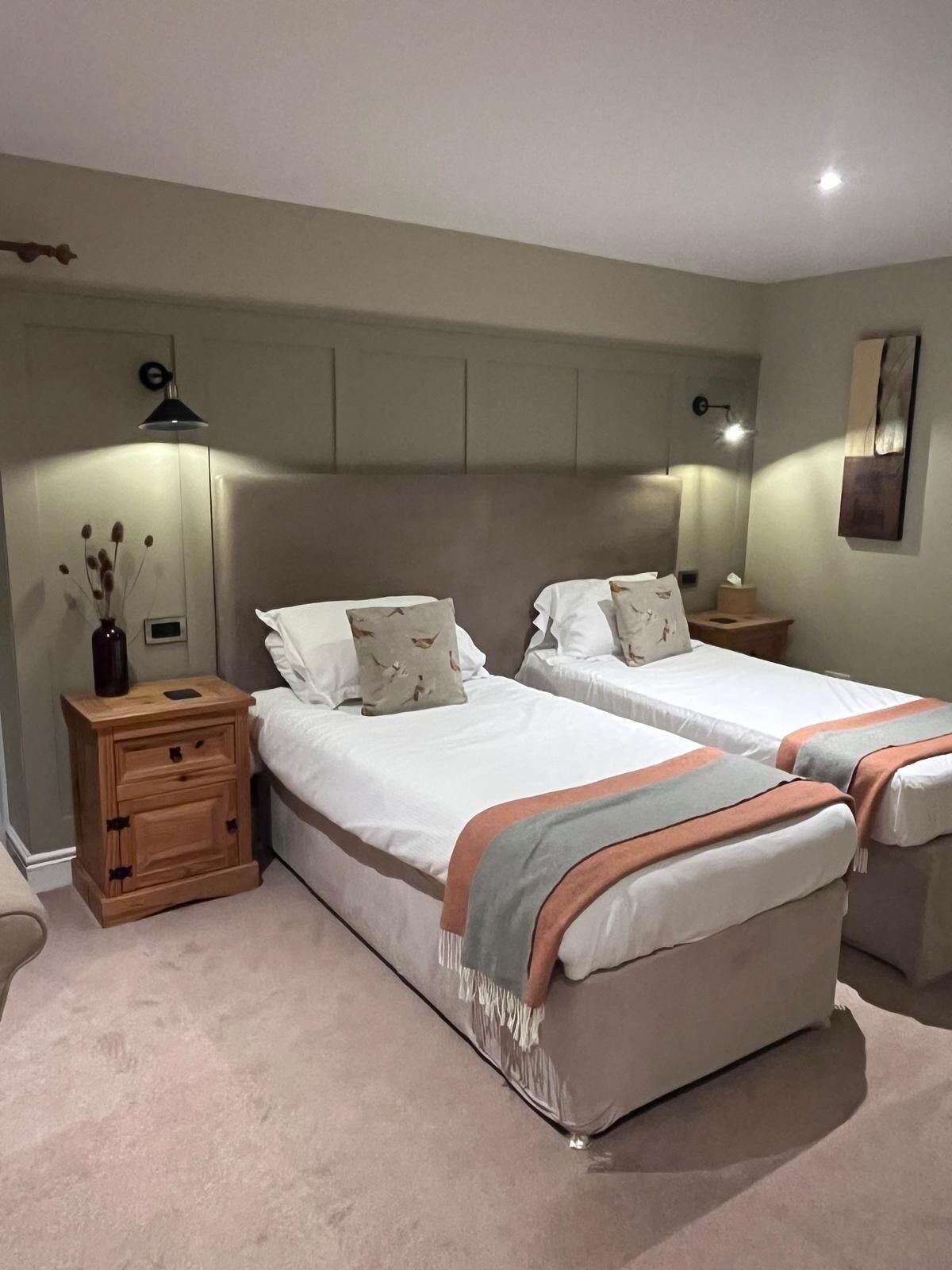 A bedroom with two beds, a nightstand, a lamp, and a wall-mounted light fixture.