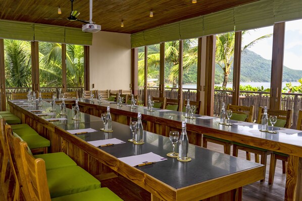 Una larga mesa de comedor con sillas, preparada para una comida, en una habitación con ventanas amplias que dan a un paisaje tropical.