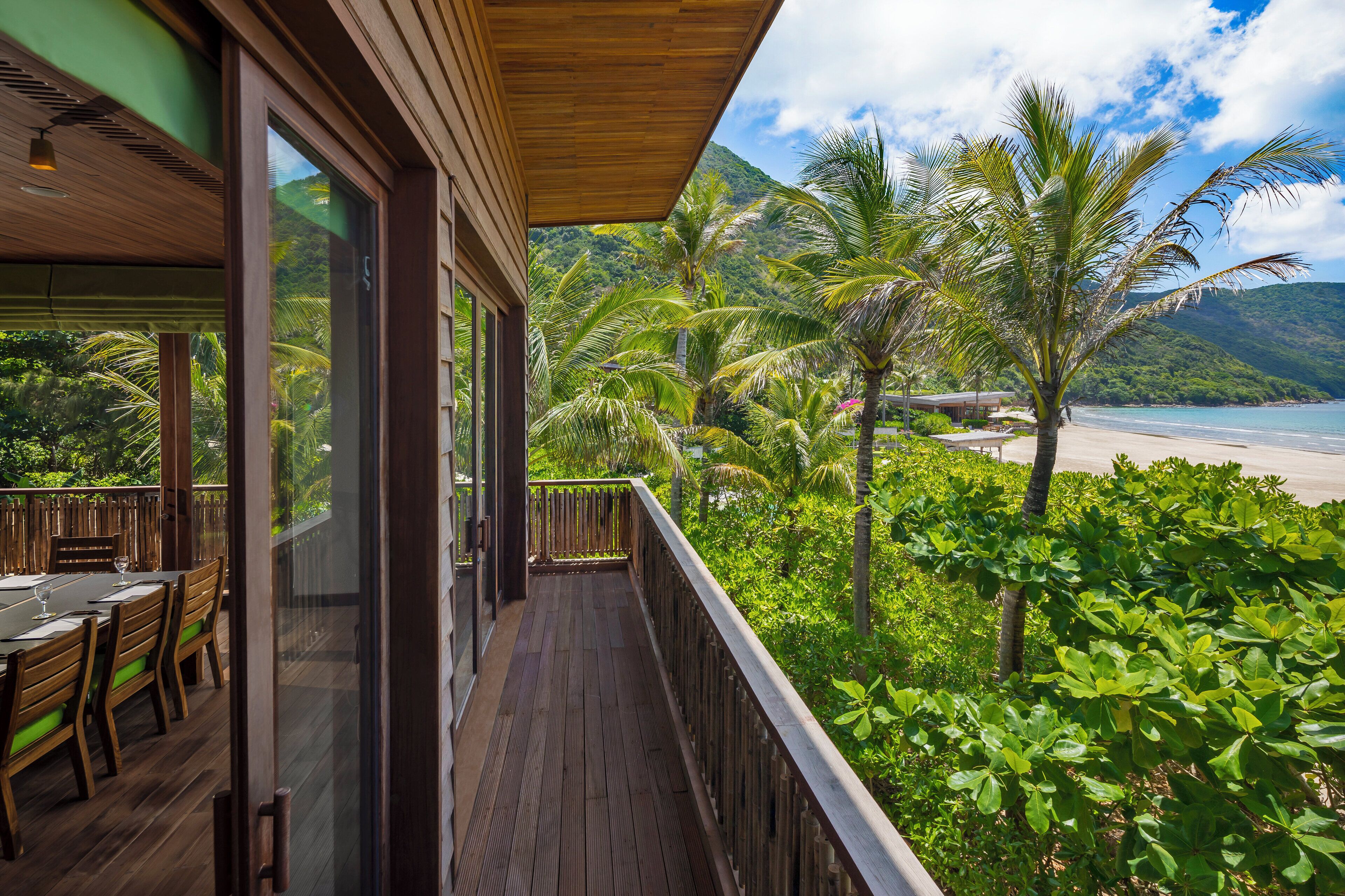A wooden deck with a glass balustrade overlooking a beach and lush greenery.