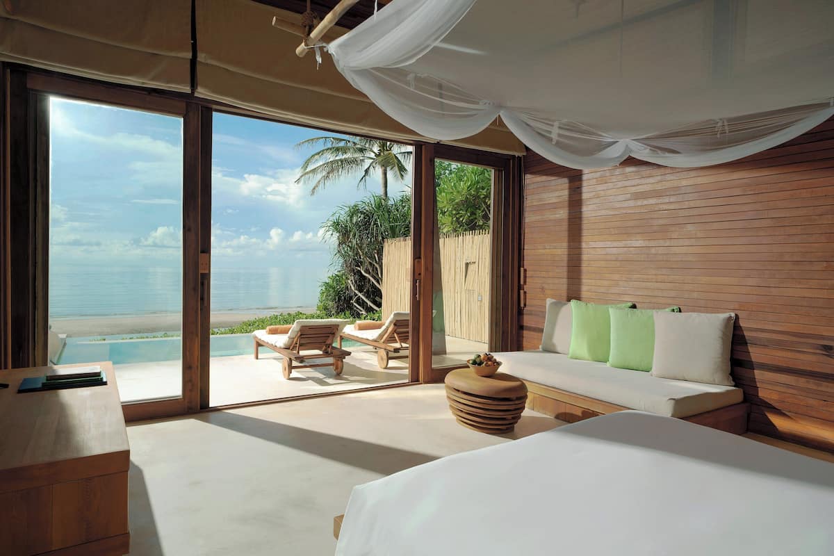 A bedroom with a large window, a bed, a wooden headboard, and a view of the beach.