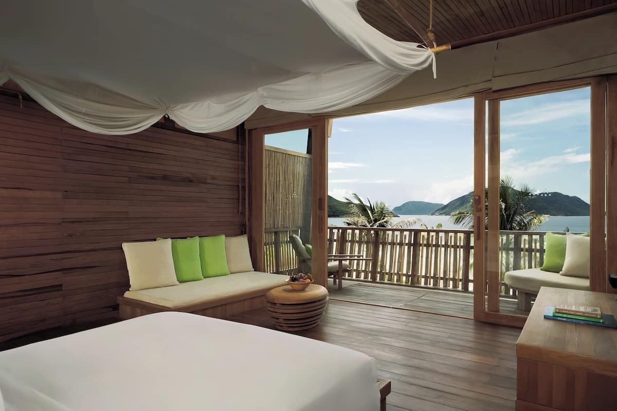 A room with a large bed, a sofa with cushions, a small round table with a bowl of fruit, and a view of the sea through large windows.