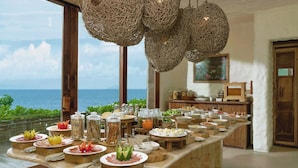 A buffet setup with various food items and condiments, a view of the ocean through large windows, and wicker hanging light fixtures. - Six Senses Con Dao (Ho Chi Minh City)