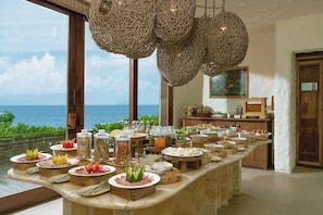 A buffet setup with various food items and condiments, a view of the ocean through large windows, and wicker hanging light fixtures. - Six Senses Con Dao (Ho Chi Minh City)