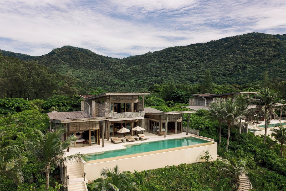 A two-story house with a swimming pool, surrounded by lush greenery and hills.