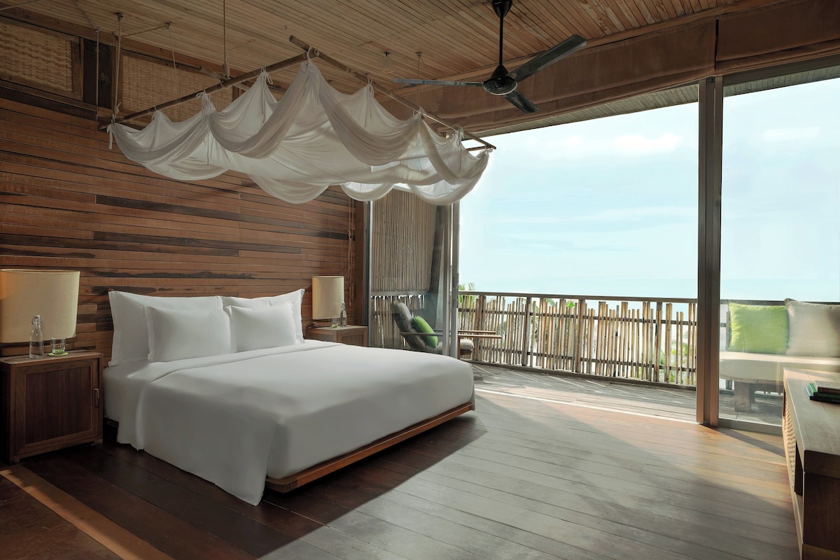 A spacious bedroom with a large bed, wooden flooring, a ceiling fan, and a balcony with a view.