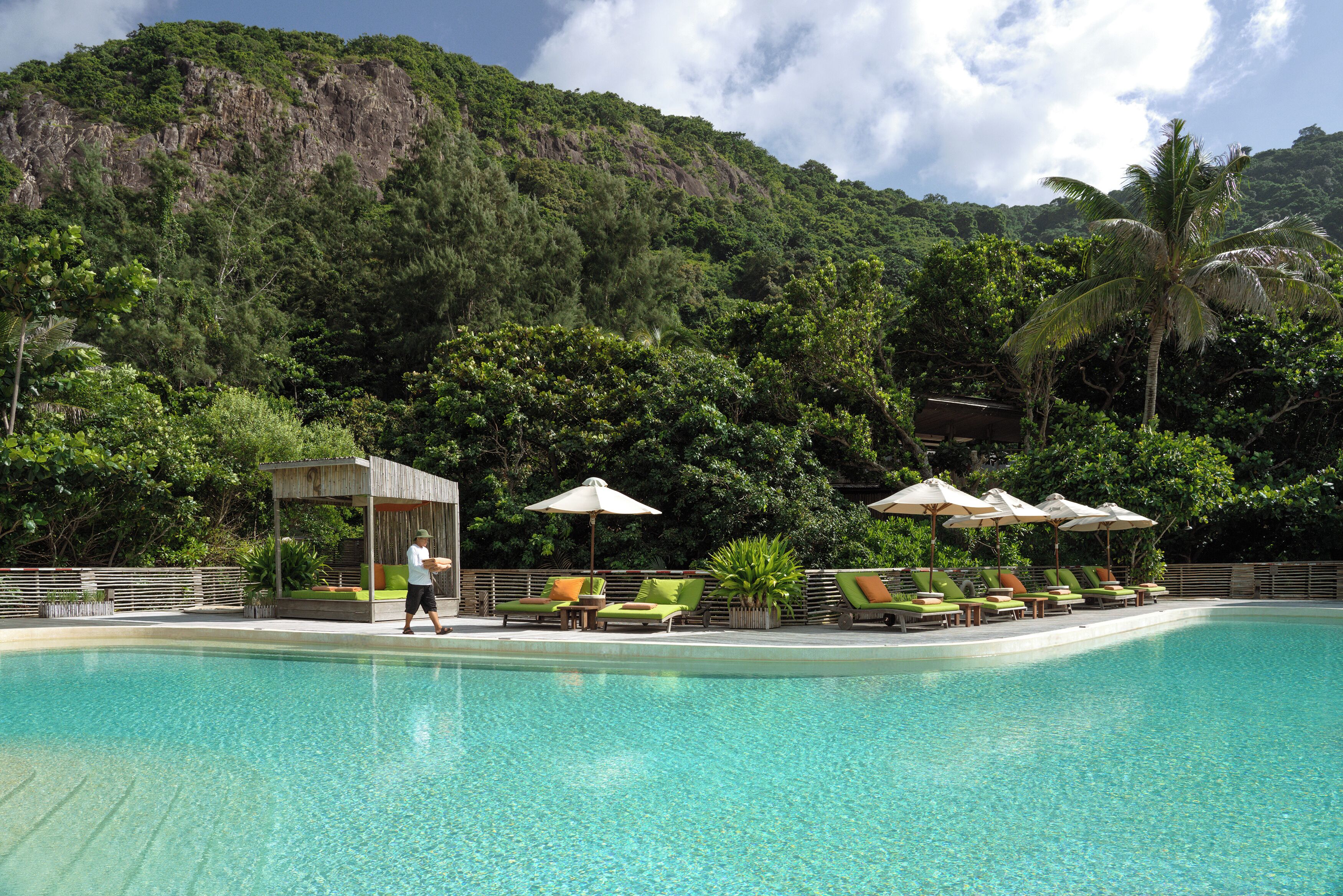 A swimming pool with lounge chairs and umbrellas, surrounded by lush greenery and a mountain backdrop.