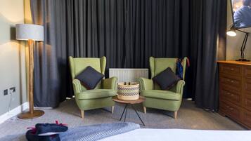 A hotel room with two green armchairs, a small round table, a basket, a bed, a dresser, and a television on the wall.