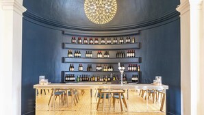 A bar area with a central circular light fixture, shelves of wine bottles, and wooden chairs around a bar counter.