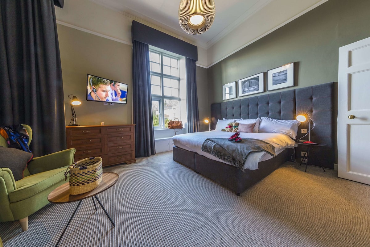 A hotel room with a large bed, a television, a desk with a lamp, a chair, and a window with curtains.