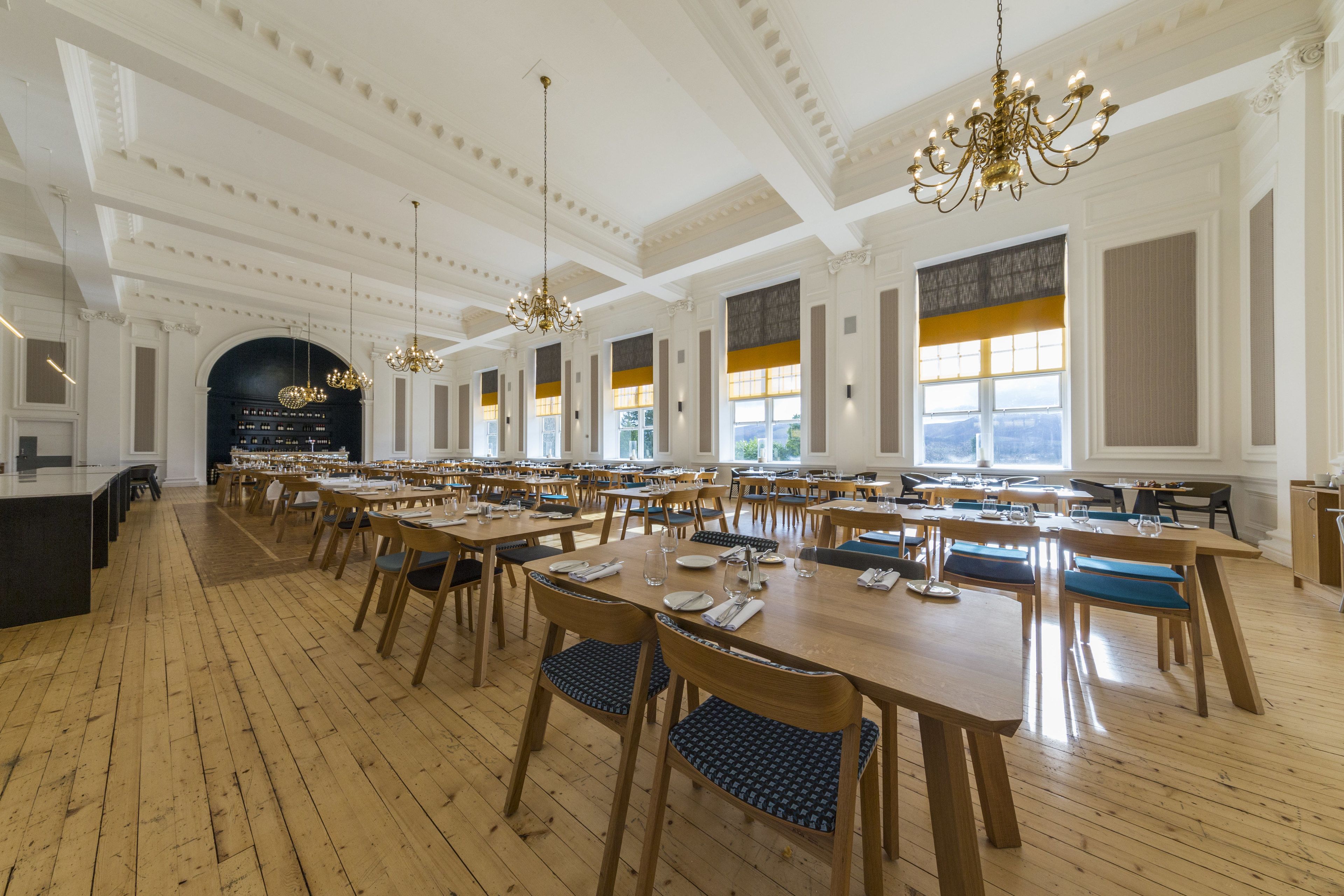 A spacious dining area with wooden tables and chairs, chandeliers, and large windows with yellow-tinted glass.