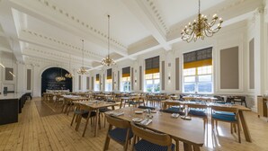 A spacious dining area with wooden tables and chairs, chandeliers, and large windows with yellow-tinted glass.