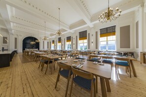 A spacious dining area with wooden tables and chairs, chandeliers, and large windows with yellow-tinted glass.