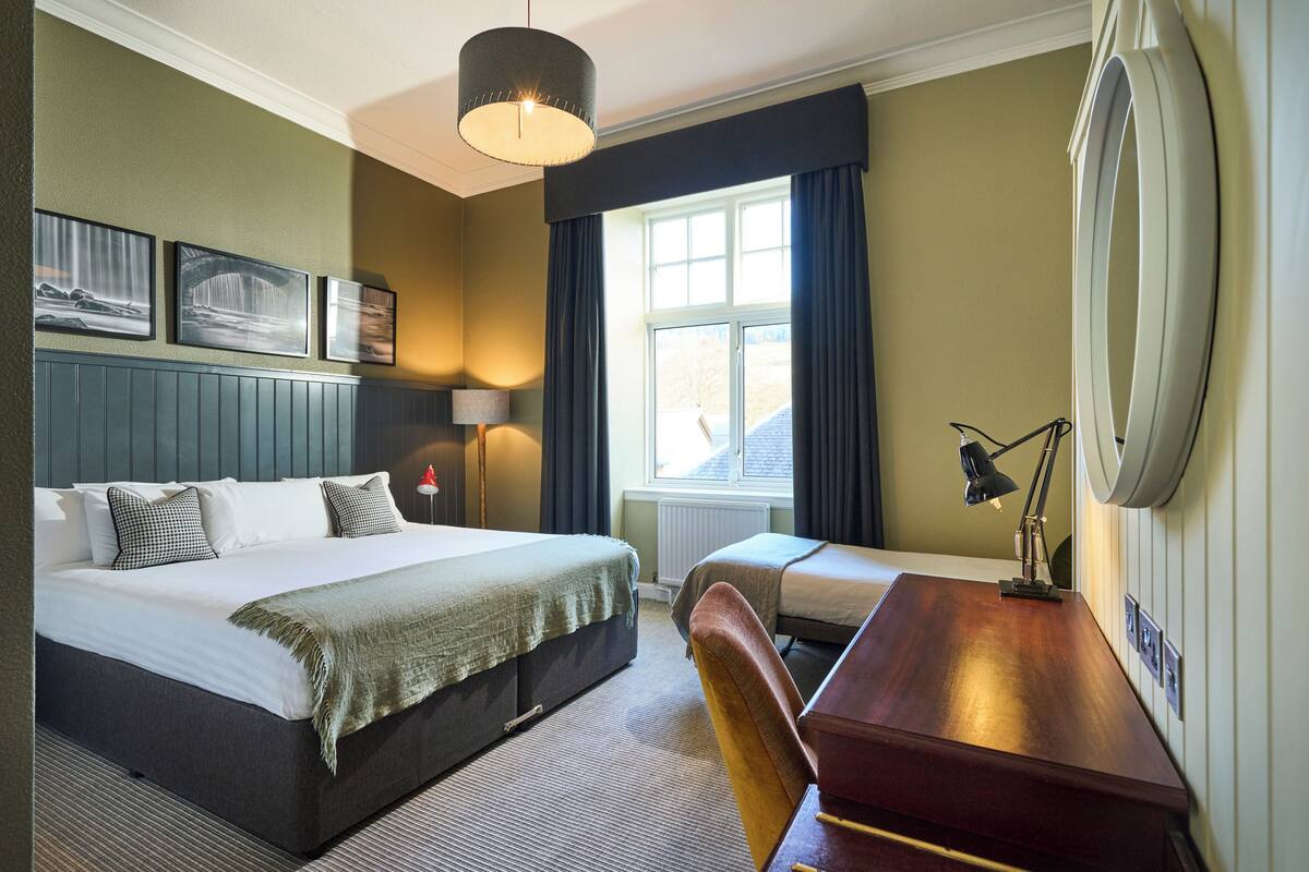 A hotel room with a large bed, a smaller bed, a desk with a chair, a lamp, and a window with curtains.