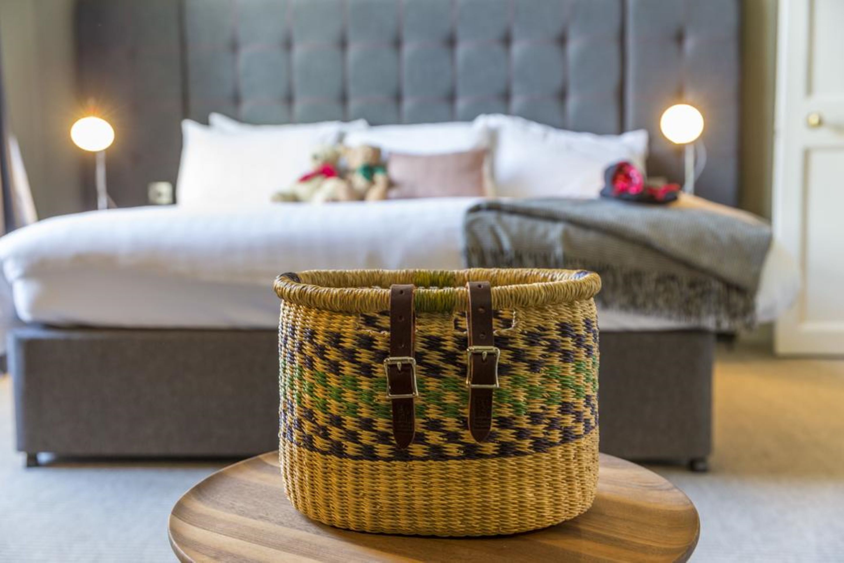 Una cesta de mimbre con correas de cuero sobre una mesa de madera en una habitación de hotel con una cama y lámparas de noche.