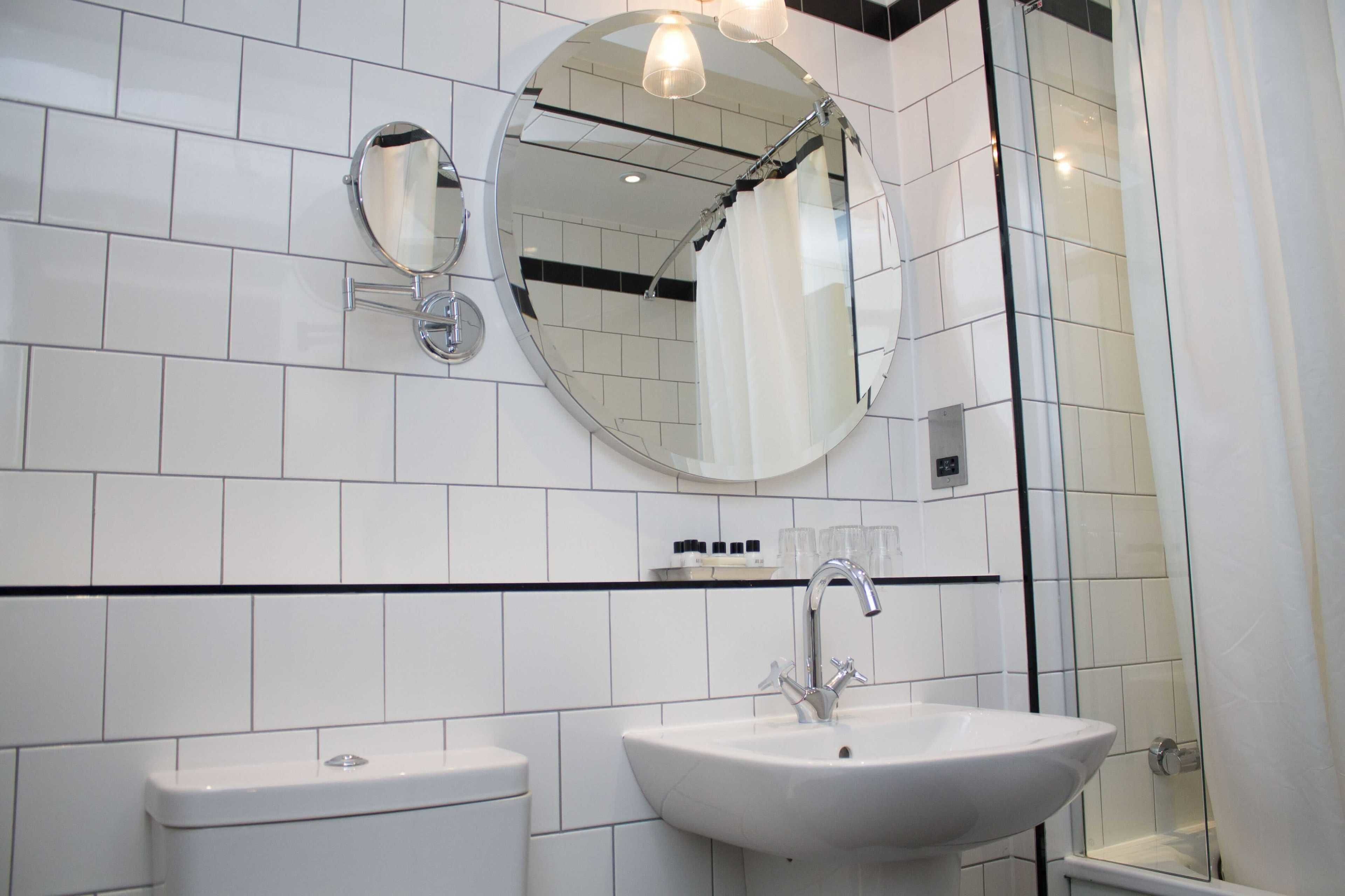 A bathroom with white tiles, a round mirror, a sink, and a shower.
