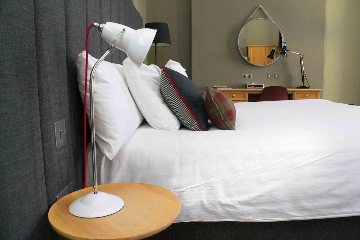A neatly made bed with white linens, a round wooden bedside table with a white lamp, a mirror above the bed, and a desk with a lamp and a red chair.