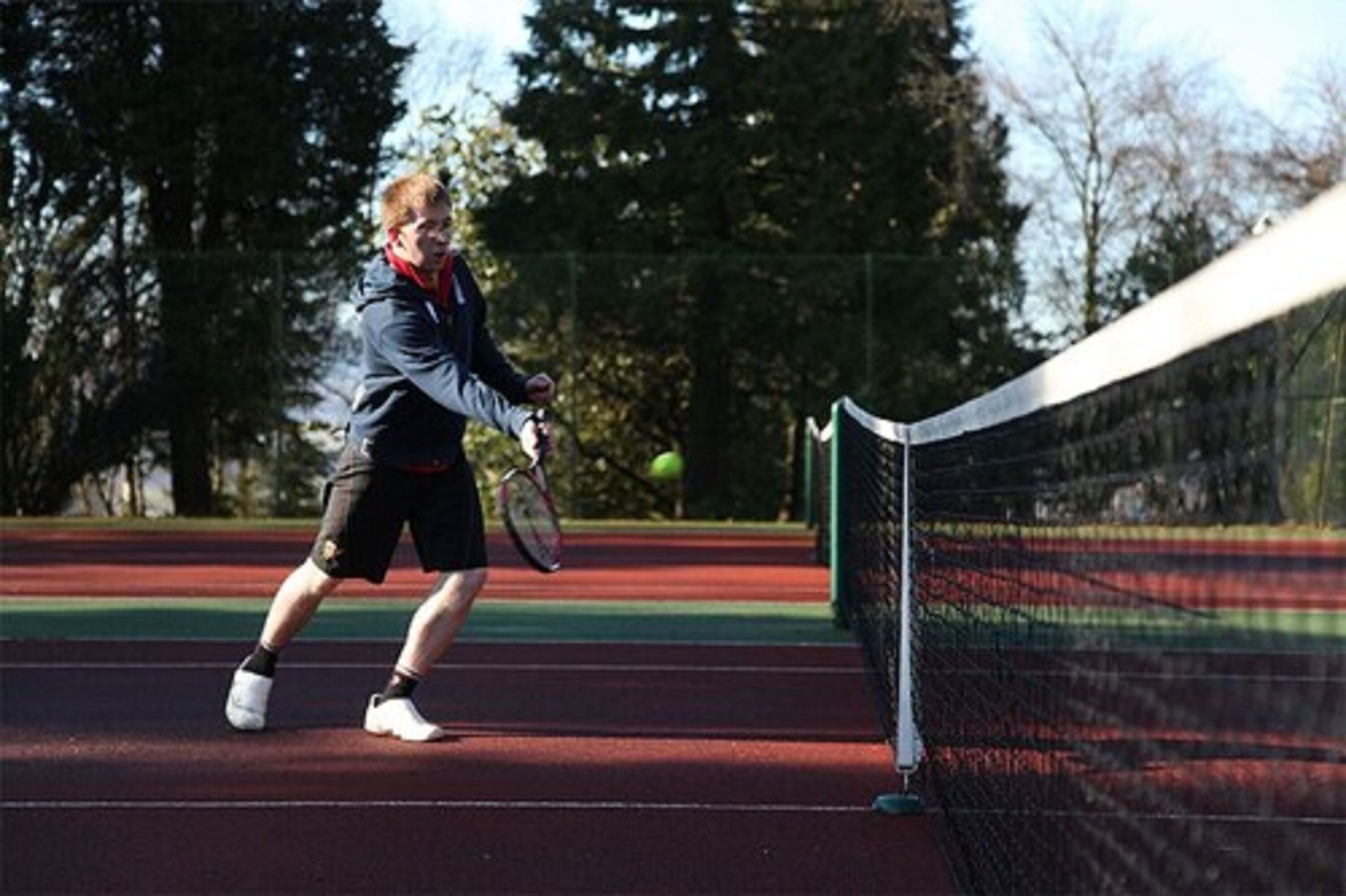 A tennis court with a net, trees, and a tennis ball in motion.