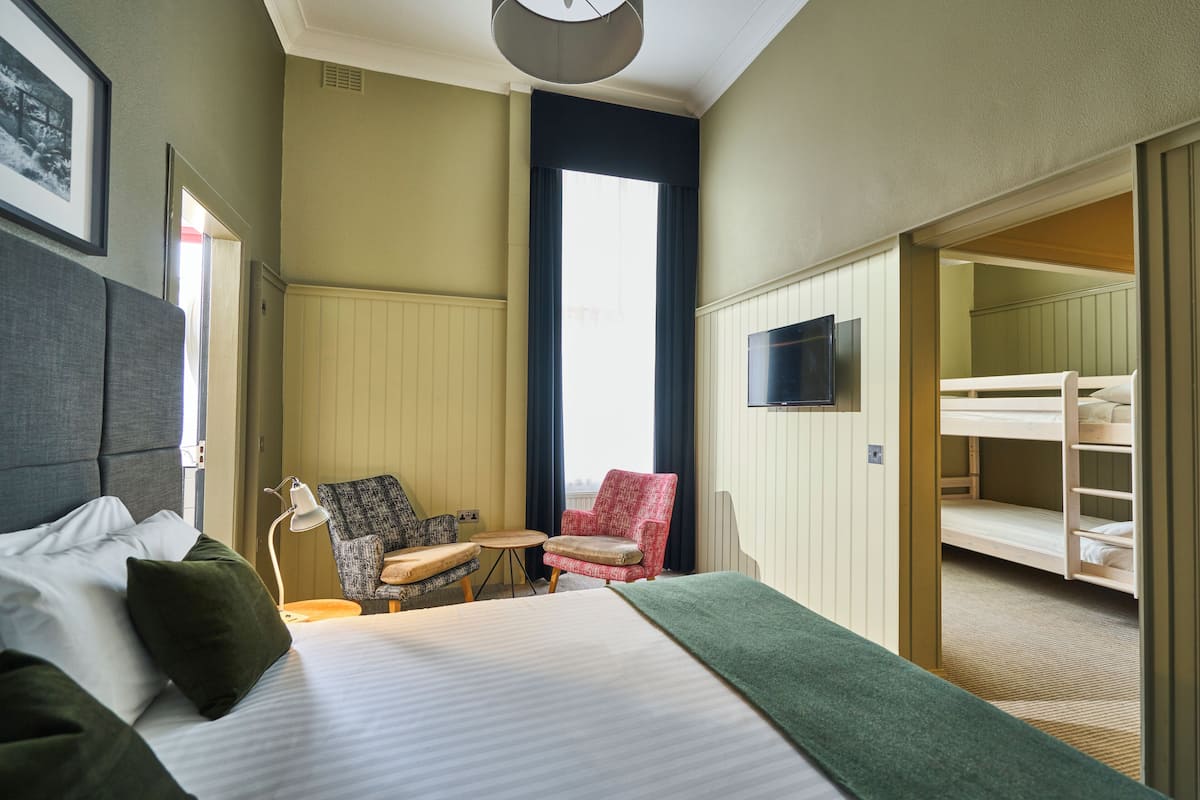 A hotel room with a bed, two chairs, a small table, a TV, and a bunk bed in the adjacent room.