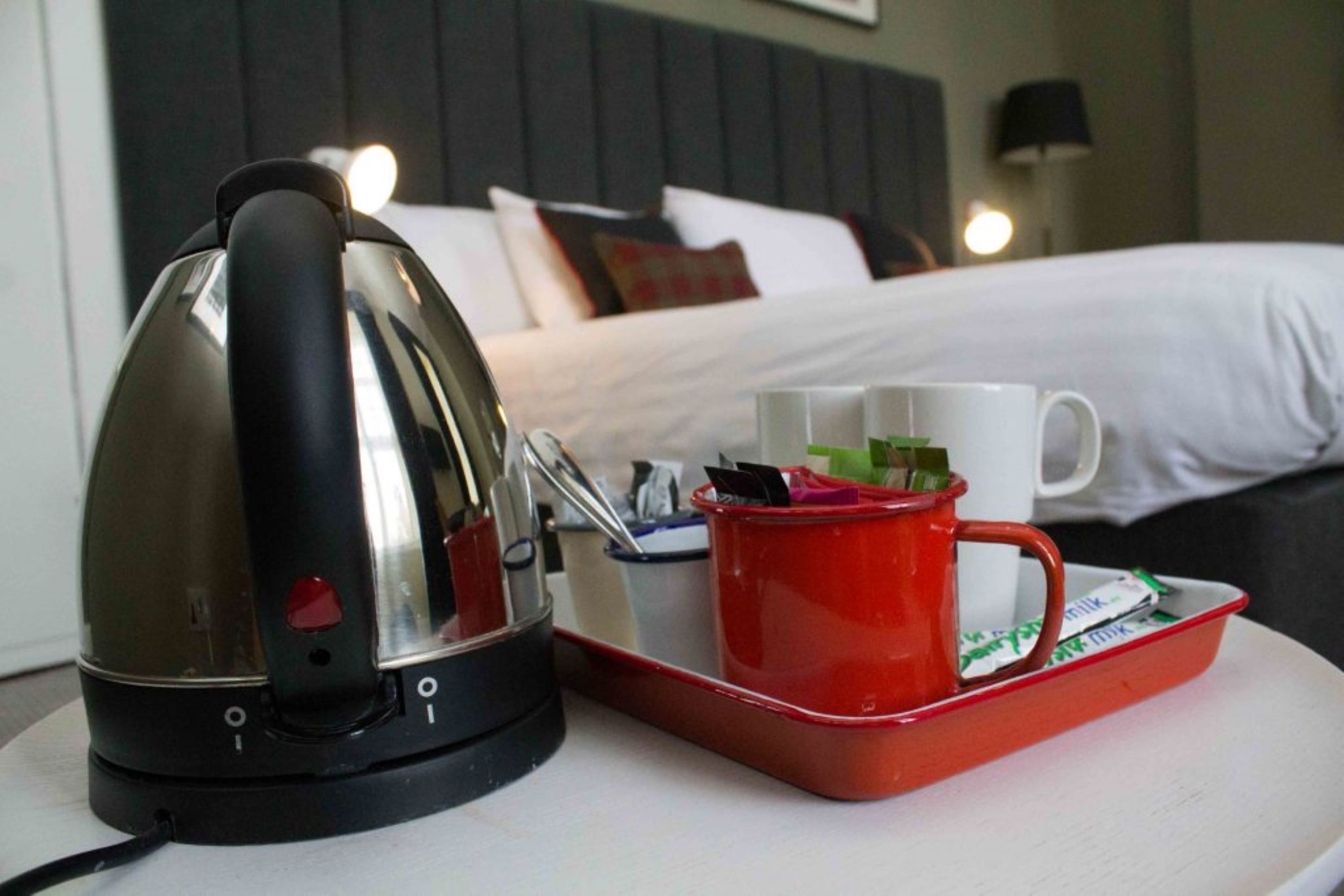 A kettle, a tray with a mug and a red cup, and a bed with pillows in a hotel room.