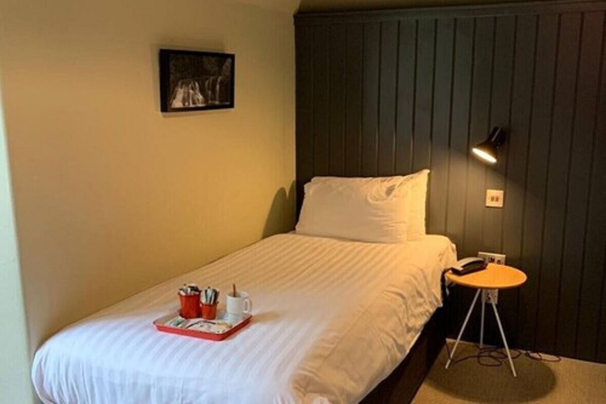 A single bed with a white comforter, a small bedside table with a lamp, a tray with a cup and a book, and a framed picture on the wall.
