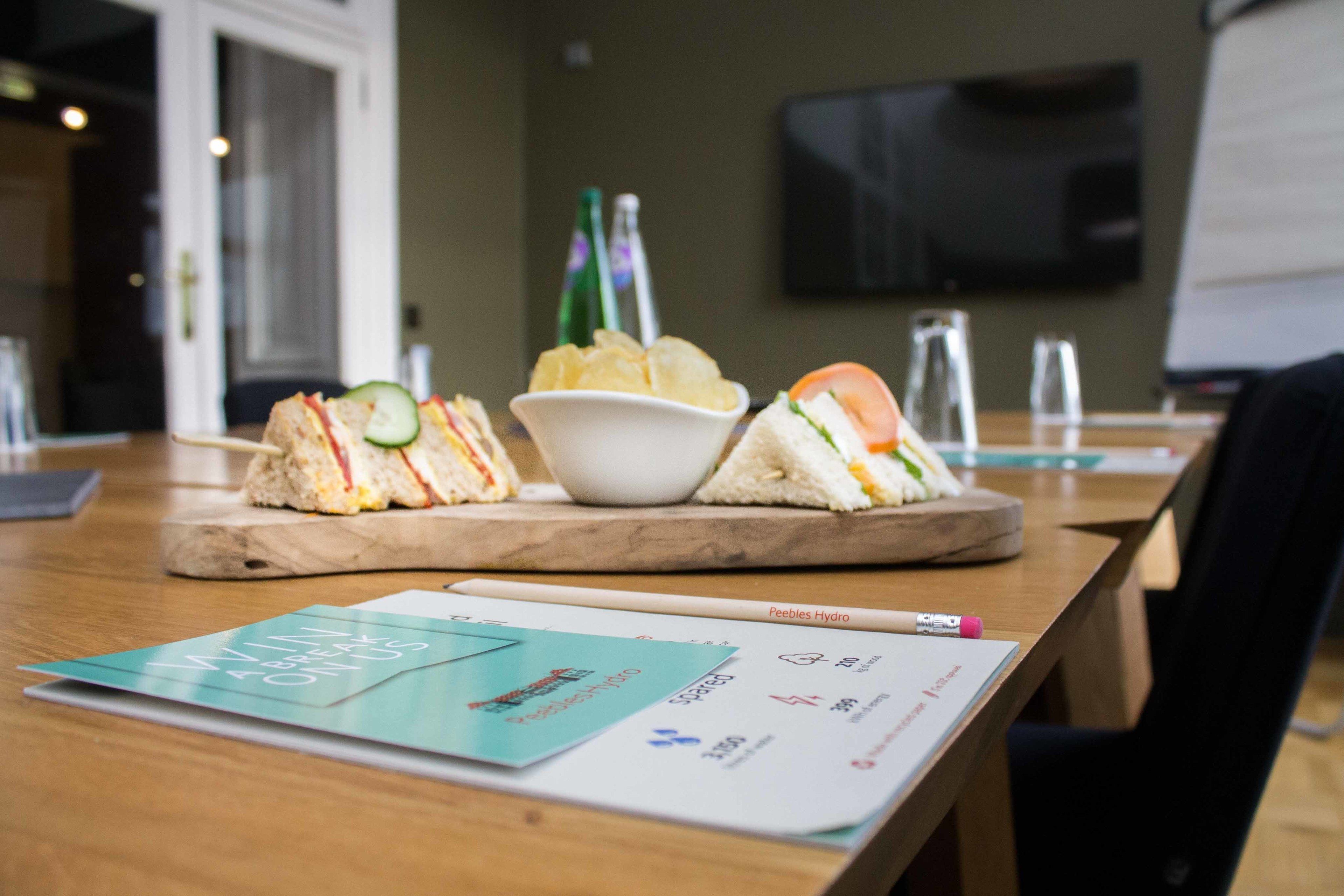 A wooden table with a tray holding sandwiches and a bowl of chips. A menu and a pen are on the table.