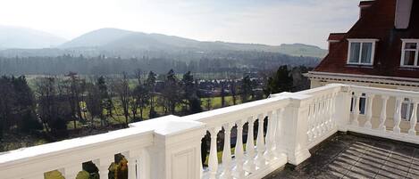 Een balkon met een witte balustrade, met uitzicht op een landschap met bergen en een stad in de verte.