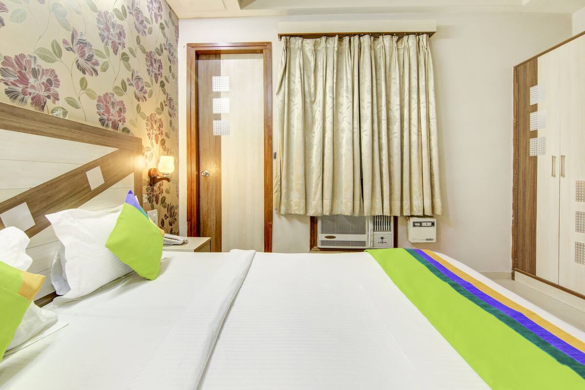 A hotel room with a bed, a wardrobe, a window with curtains, a bedside table, and a wall-mounted air conditioner.