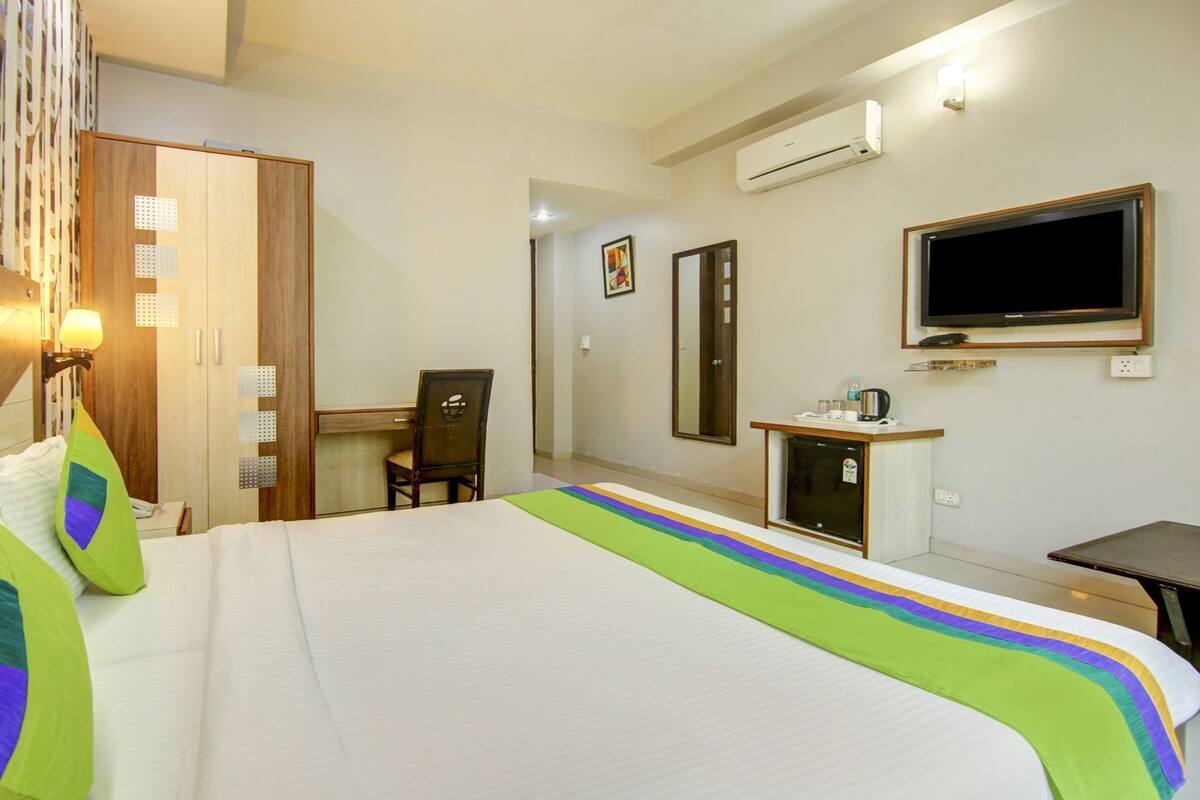 A hotel room with a bed, a desk, a chair, a TV, and an air conditioning unit.