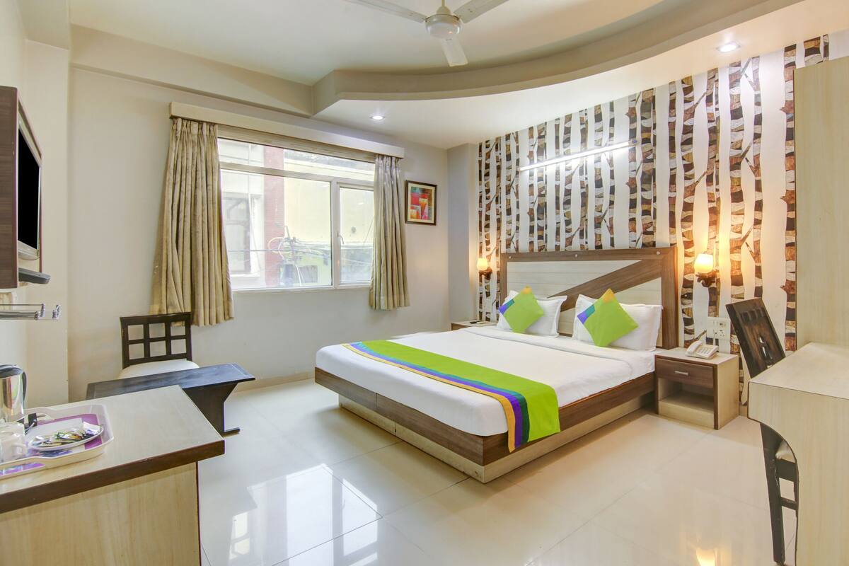 A hotel room with a bed, a desk, a chair, a window with curtains, a wall with a patterned design, and a ceiling fan.