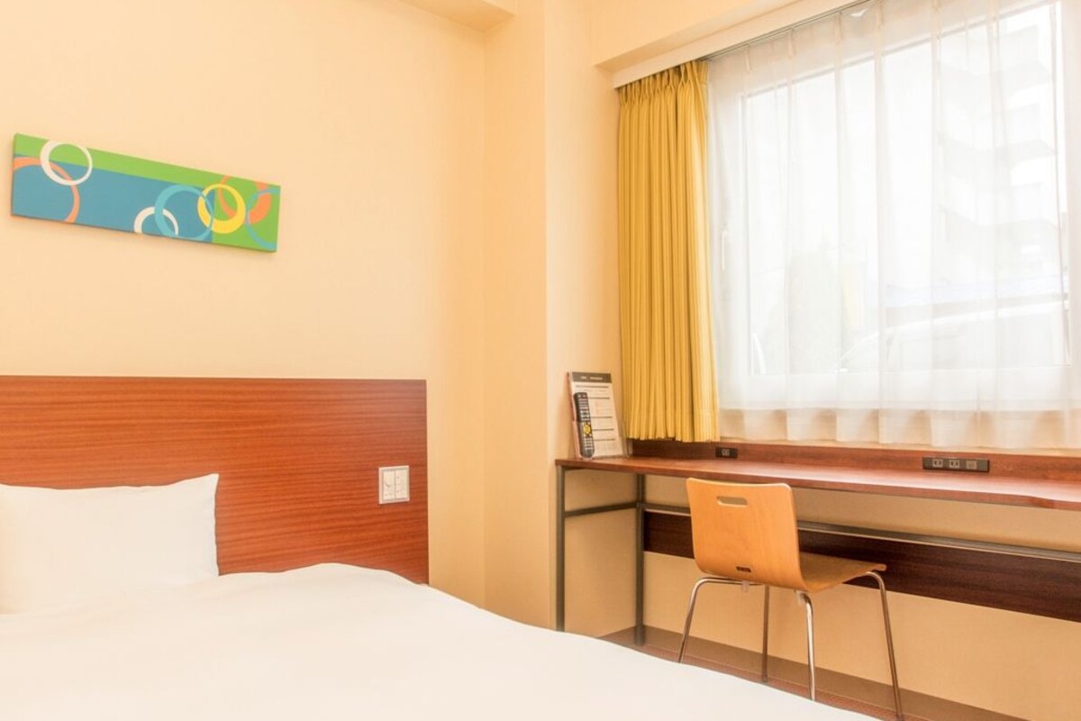 Hotelkamer met een bed, bureau, stoel, raam met gordijnen en een kleurrijk kunstwerk aan de muur.