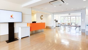 A modern hotel lobby with a reception desk, a large screen displaying the Comfort logo, and a dining area with chairs and tables.