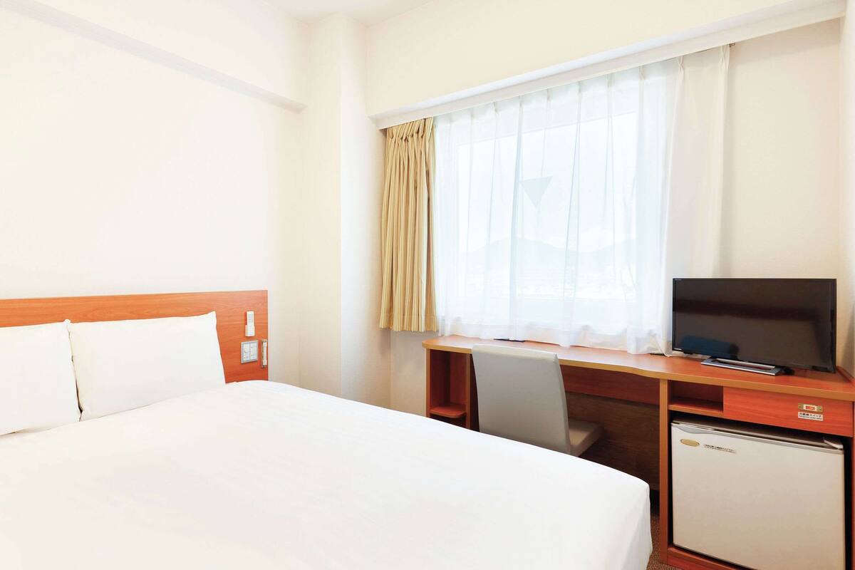 A hotel room with a bed, a desk with a television, a small refrigerator, and a window with curtains.