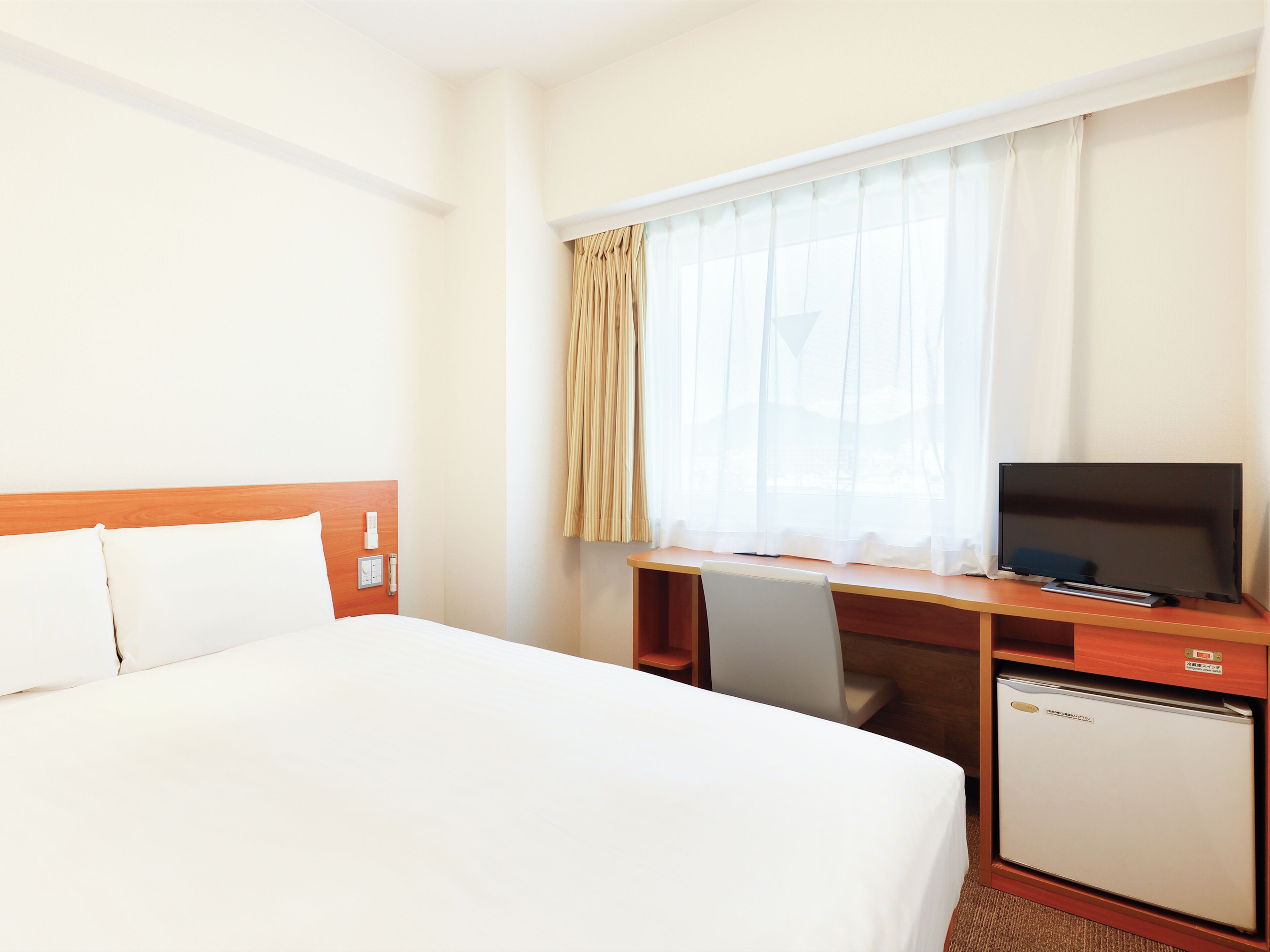 Une chambre d’hôtel avec un lit, un bureau avec une télévision, un petit réfrigérateur et une fenêtre avec des rideaux.