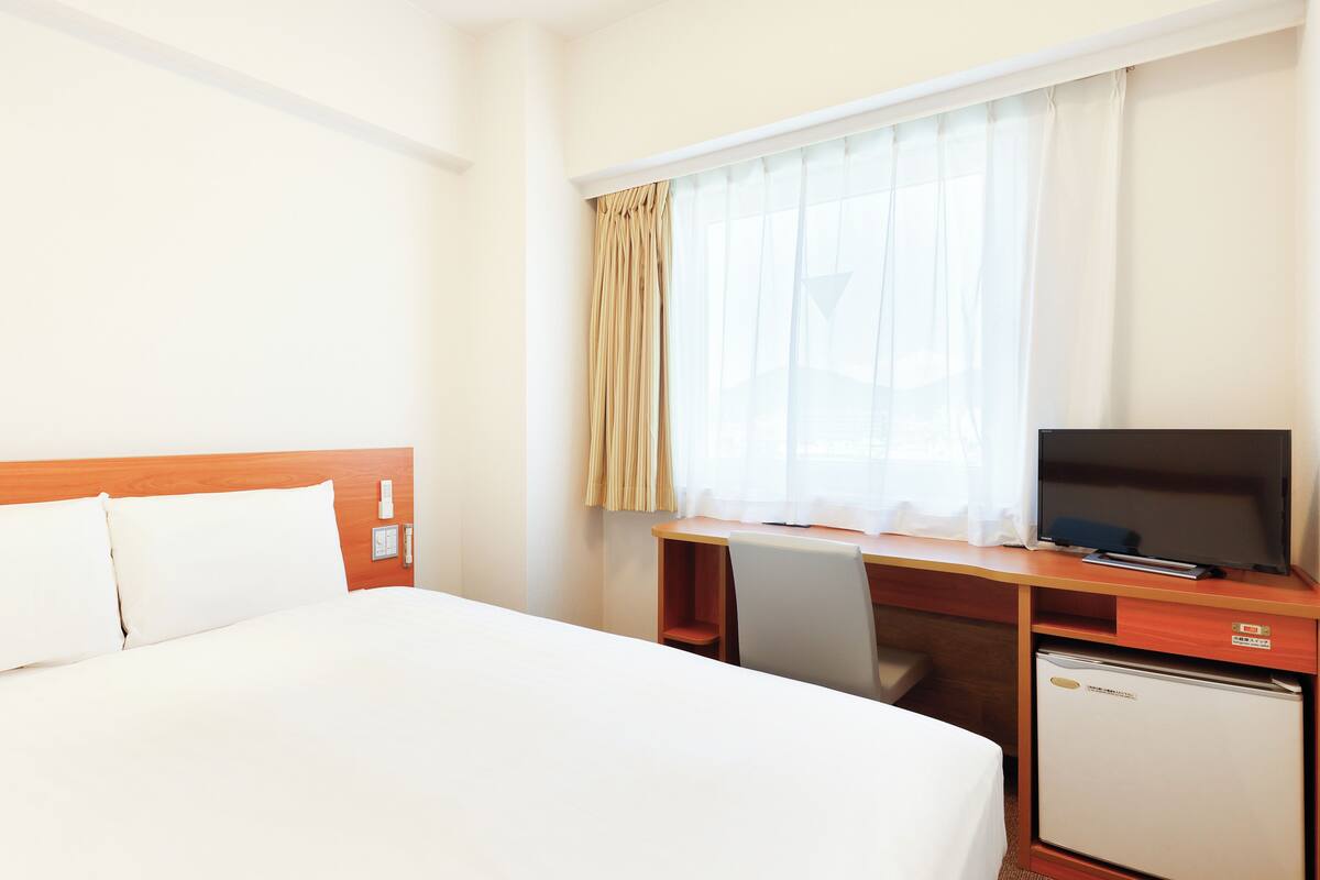 A hotel room with a bed, a desk with a television, a small refrigerator, and a window with curtains.