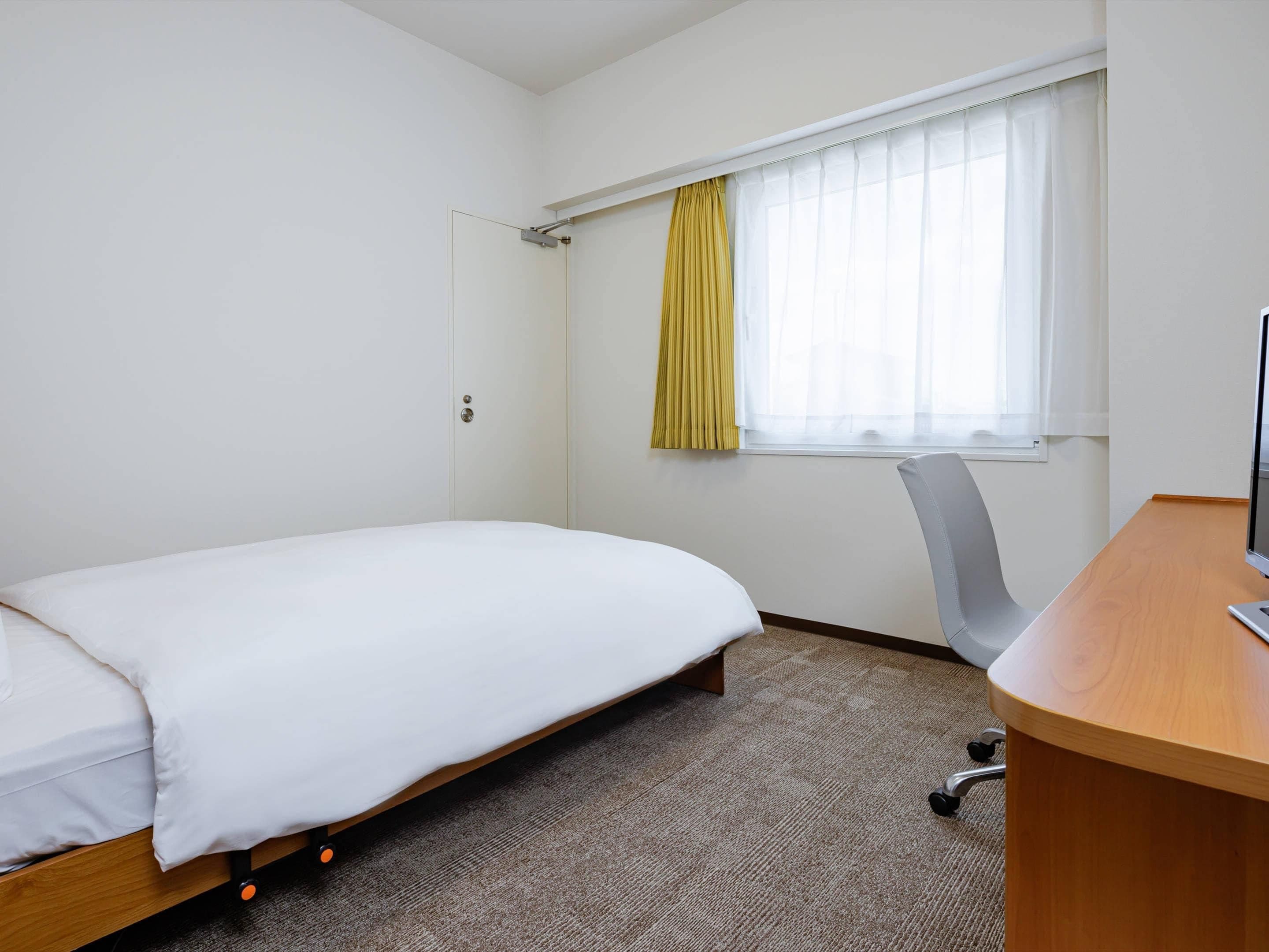 A hotel room with a single bed, a desk with a chair, a window with curtains, and a door.