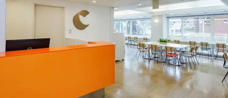 Una moderna zona de recepción con un mostrador naranja, una gran decoración mural en forma de C y un comedor con mesas y sillas.