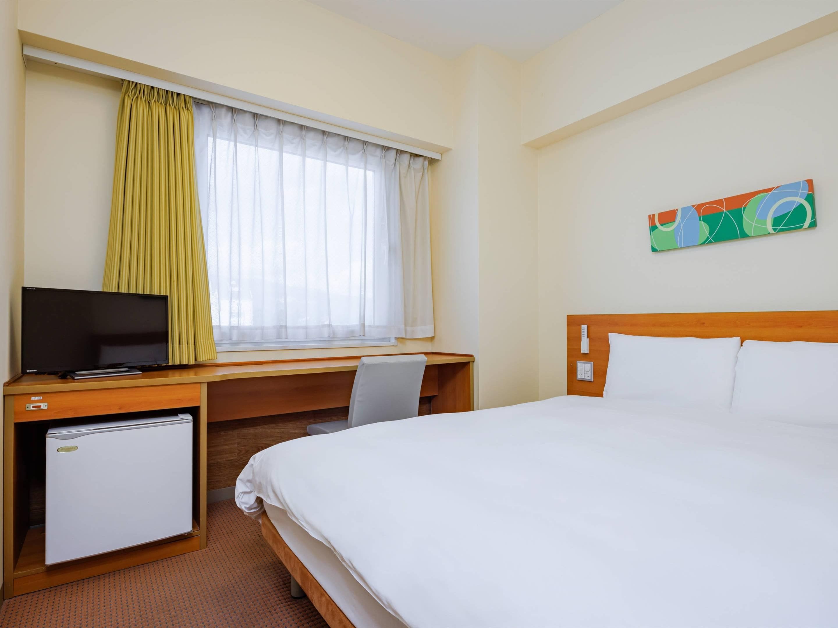 A hotel room with a bed, a desk with a television, a small refrigerator, a window with curtains, and a wall-mounted painting.