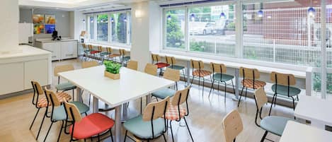 A spacious dining area with multiple tables and chairs, large windows with a view of the outside, and a kitchen area in the background.