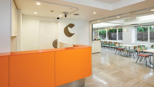 Un espace d’accueil au design moderne, avec un comptoir orange vif, une grande lettre « C » fixée au mur et une vue sur un espace de restauration avec des tables et des chaises.